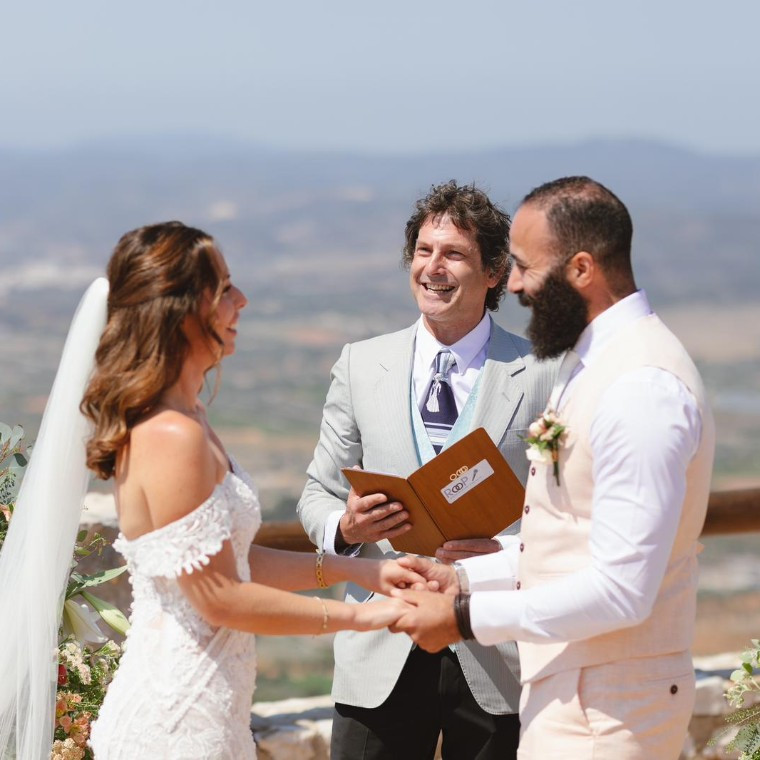 Bride and groom holding hands during an outdoor wedding ceremony with their celebrant, surrounded by flowers overlooking the countryside.