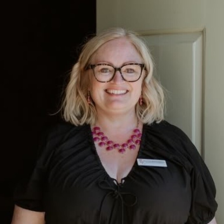 Ruth standing in front of a door after conducting a ceremony.  Ruth is smiling, wearing a black dress with a pink necklace.