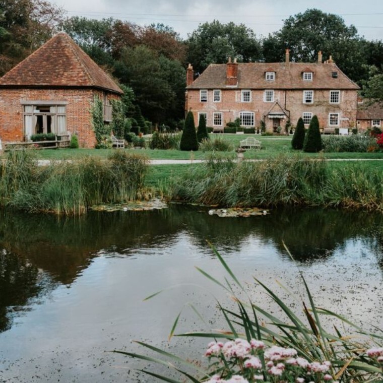 A tranquil view of Solton Manor in Kent, showing the historic red-brick dovecote and the Grade II-listed Manor House reflected in a large wildlife pond, surrounded by lush gardens, meadow grasses and 
