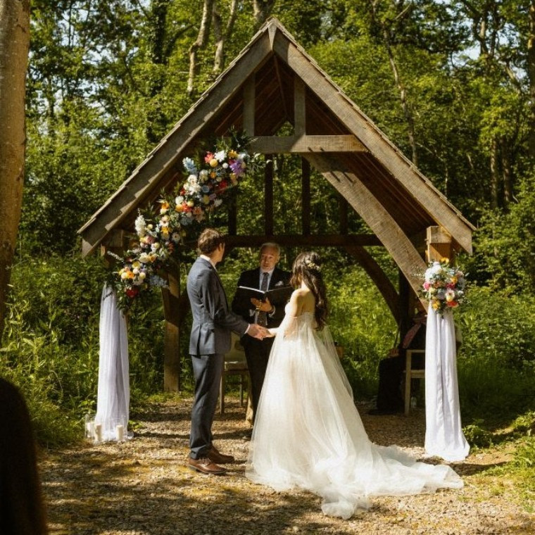 A bride and groom stand underneath an oak arbour in a woodland in front of a celebrant