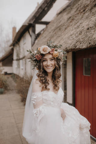 bride with a large flower crown standing in front of a cottage smiling at the camera