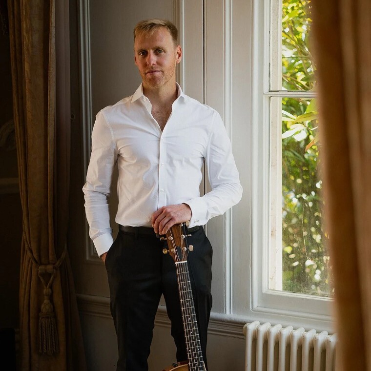 Matt Rayner, an Essex-based wedding musician, standing by a window, holding an acoustic guitar.