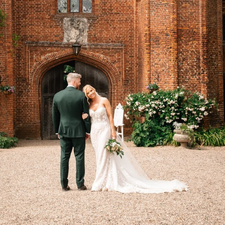 Bride and groom arm in arm outside the red-brick Tudor gatehouse at Leez Priory, Essex, the bride smiling at the camera – wedding photography by Pixton Media, Telford