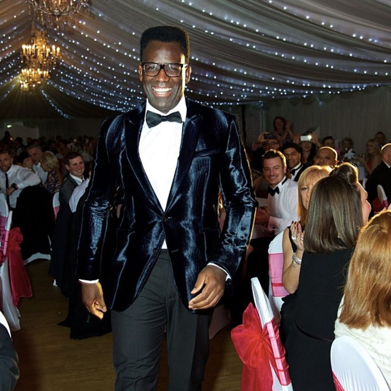 Terry J Lewis, wedding DJ and MC, smiling in a velvet tuxedo as he walks through a marquee wedding reception full of guests under chandeliers and fairy lights