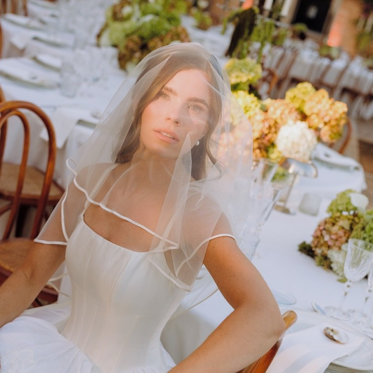 Bride beneath a sheer veil, seated beside a flower-filled wedding table in an editorial bridal portrait.