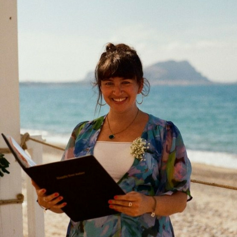 An image of a woman holding a book open, stood in front of a beach
