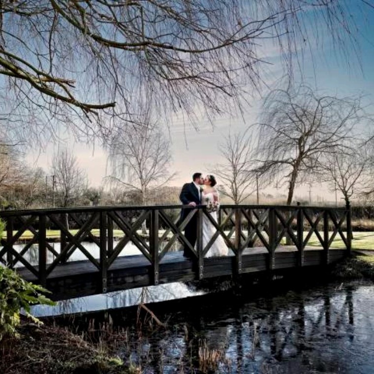 Bride and groom on wooden bridge over lake with trees around.