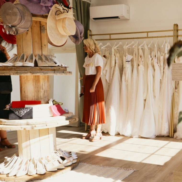 A view inside a bright, modern bridal boutique with light wooden floors. Racks of white wedding dresses are visible in the background. In the foreground, a display of hats and shoes for mothers of the