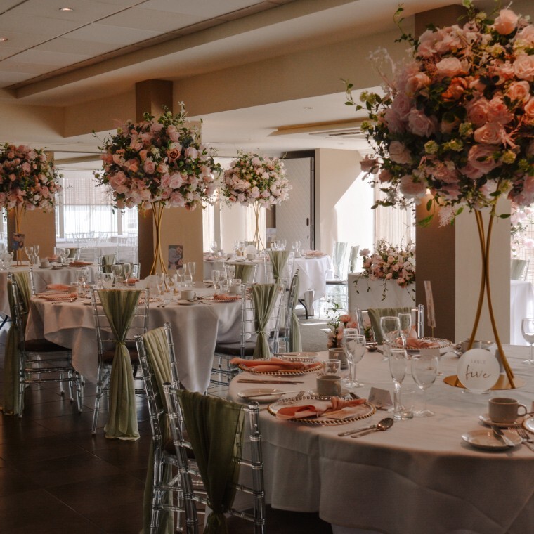 Wedding room with round wedding tables set in a banquet layout, dressed with pink napkins and sage sashes. The table centrepieces are a large spray of pink flowers with green foliage. 