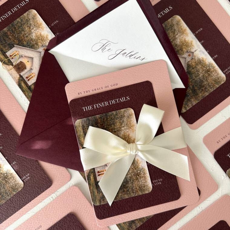 A burgundy and pink wedding invitation suite with a ribbon bow and printed envelopes