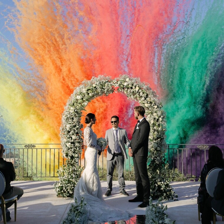 The wedding celebrant Claudio Ceino joyfully looks at the newlywed couple while colorful smoke trails cross the background of the scene.