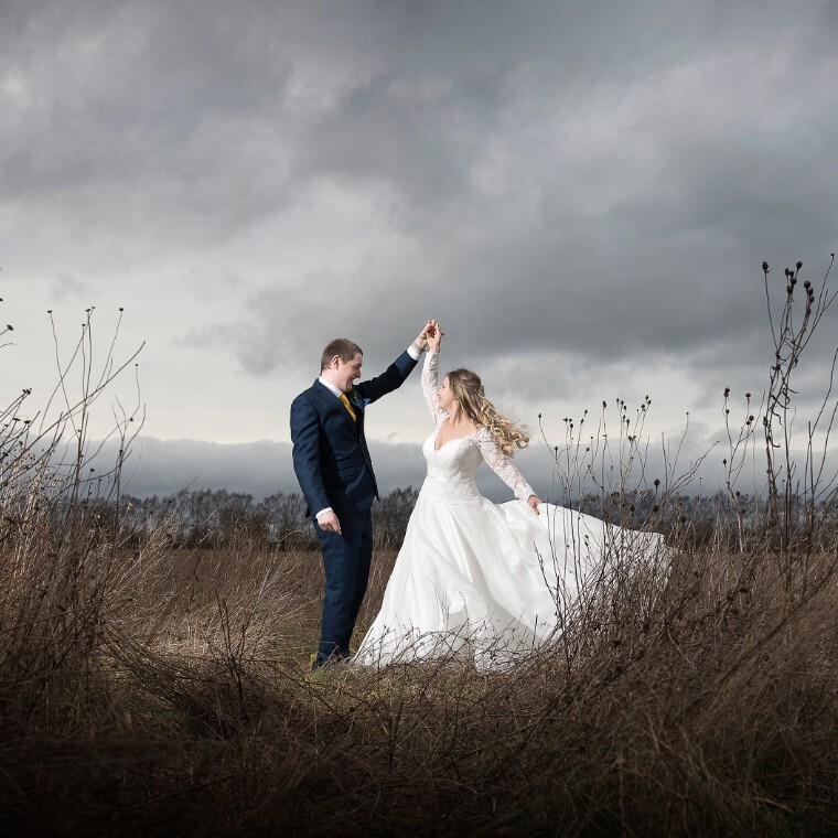 Bride in white with long blonde hair holds her dress while dancing with groom in a blue suit. They are smiling at each other, they are in a field and behind them is a grey stormy sky