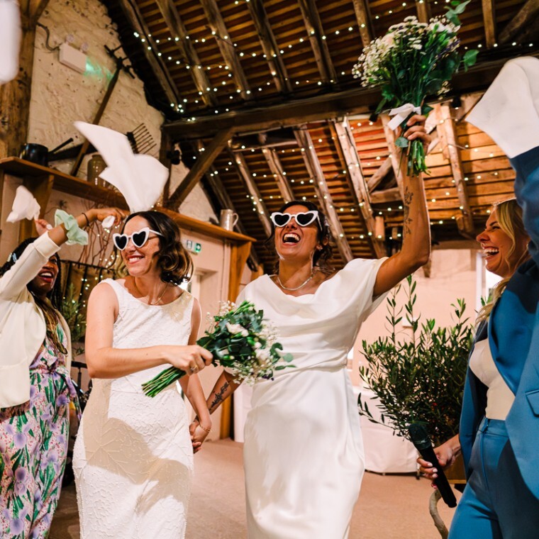 Two brides cheer as they enter their reception venue wearing loveheart sunglasses