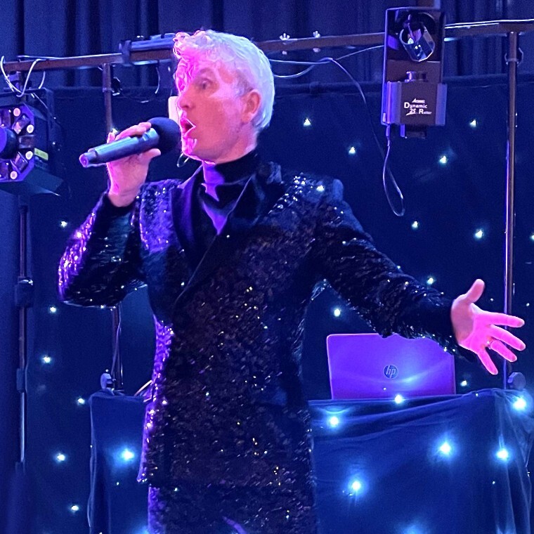 Wedding singer Peter Stanford is performing live on stage at a wedding reception. He is wearing a black sequinned suit, which is sparkling in the spotlight.