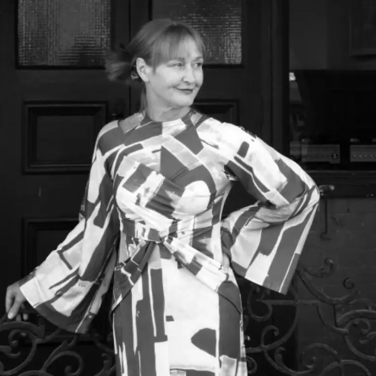 Black and white photo of a blonde woman in a graphic print dress looking away from the camera. The woman is blonde and in her 50s her hair is loosely tied up.