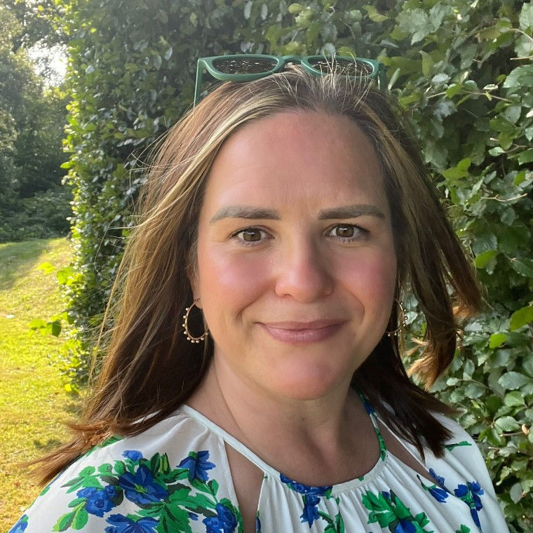 Emily Ayres smiling at the camera dressed in a while, green, and blue floral dress before a wedding ceremony