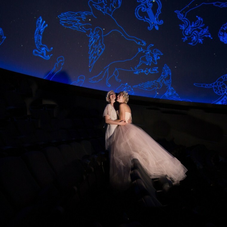 Two brides embrace in a planetarium, standing among rows of seats beneath a dome lit with glowing blue constellation artwork.