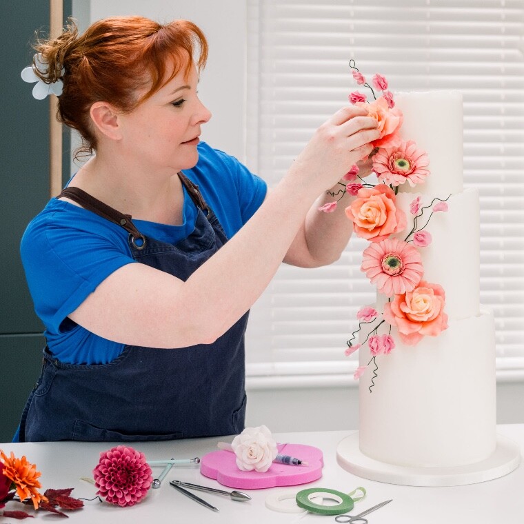 Amy from Perfect Moment Cake Design is seen setting up a tall three tier wedding cake. She is adding hand crated sugar flowers. 