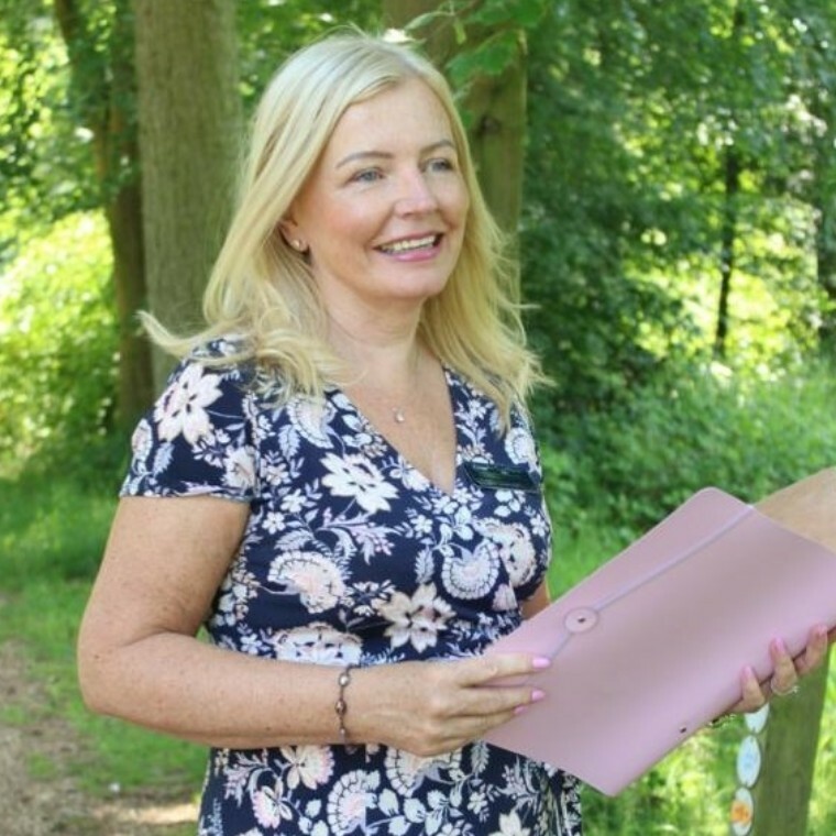 Angela Davidson, a smiling wedding celebrant, holding a ceremony folder while standing in a woodland setting