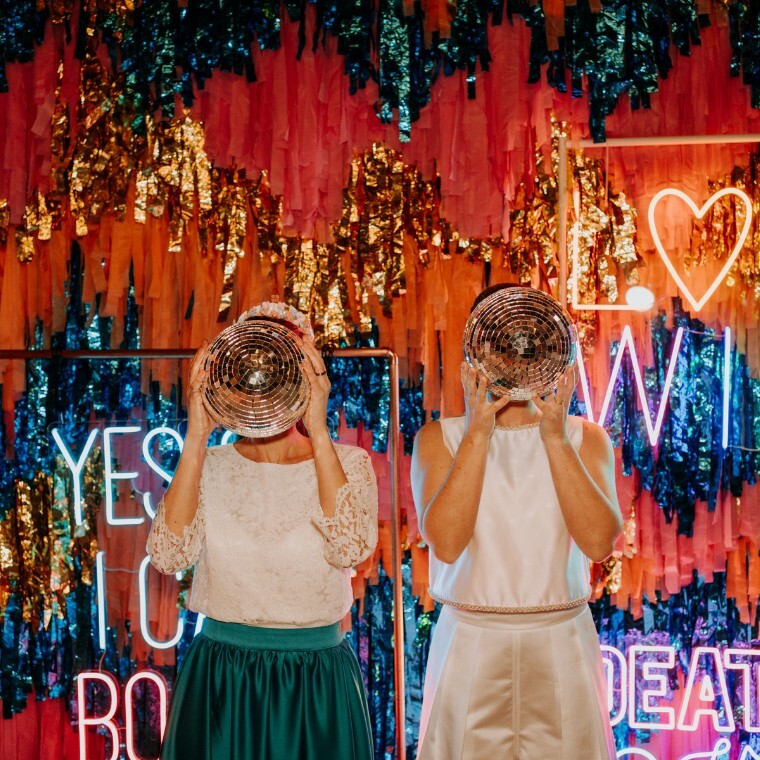 Two brides holding disco balls in front of their faces, standing in front of a vibrant, colourful fringe and neon wedding backdrop with signs reading “YES I DO” and “LOVE WINS”. Fun, bold wedding vibe