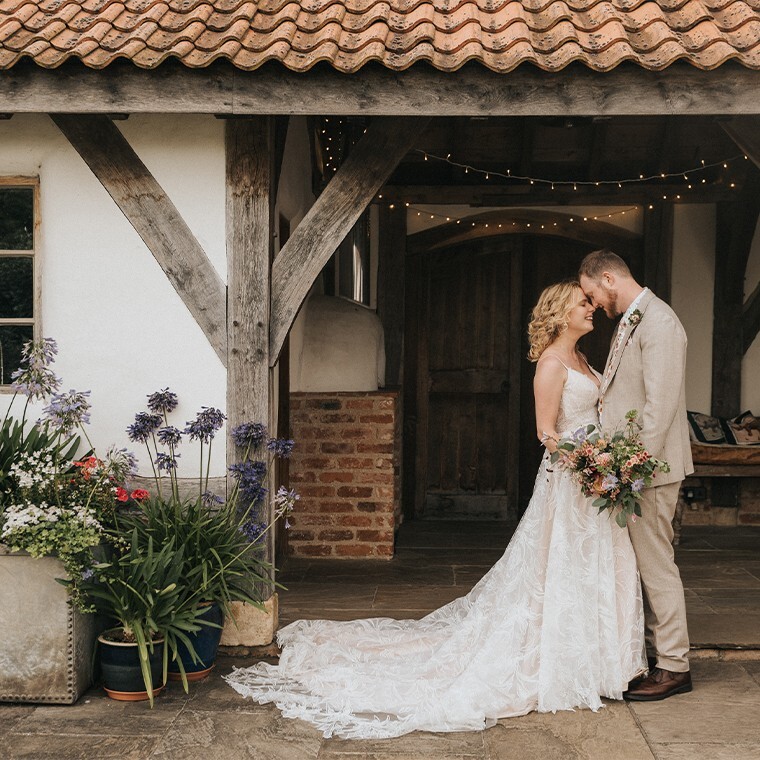 A smiling bride and groom forehead-to-forehead in front of a rustic cottage barn with fairy lights and wildflowers.