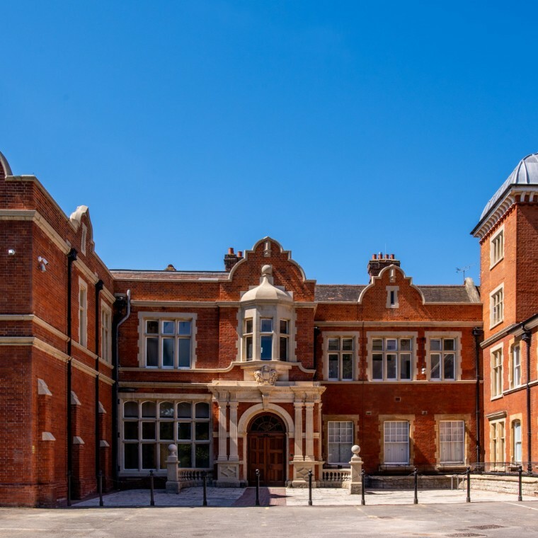 Historic brick building with ornate central entrance, white-framed windows, decorative stonework, and a prominent bay window above the door. The roofline features chimneys and gables, set against a cl